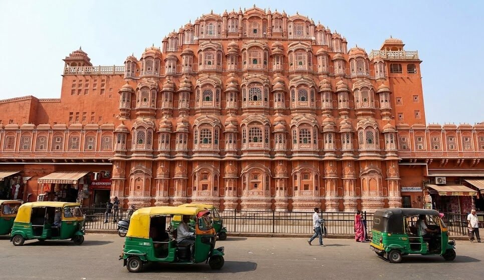 Noida to Jaipur Taxi Service with Udaipur Tour Options a street view of the hawa mahal, the palace of winds, a famous red sandstone monument in jaipur, with auto rickshaws driving past.