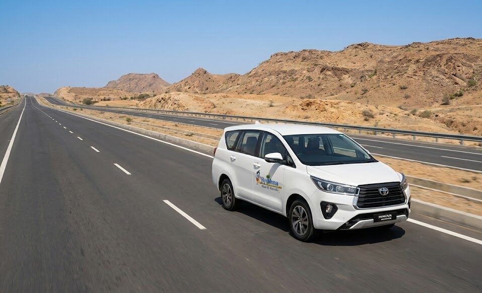 a white toyota innova crysta from shri krishna tour & travels driving on a multi lane highway through an arid, rocky rajasthan landscape.