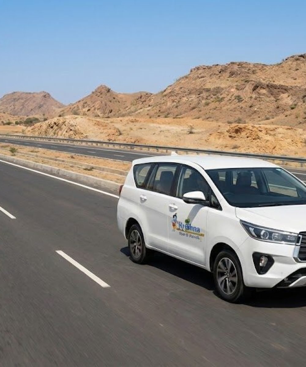 a white toyota innova crysta from shri krishna tour & travels driving on a multi lane highway through an arid, rocky rajasthan landscape.